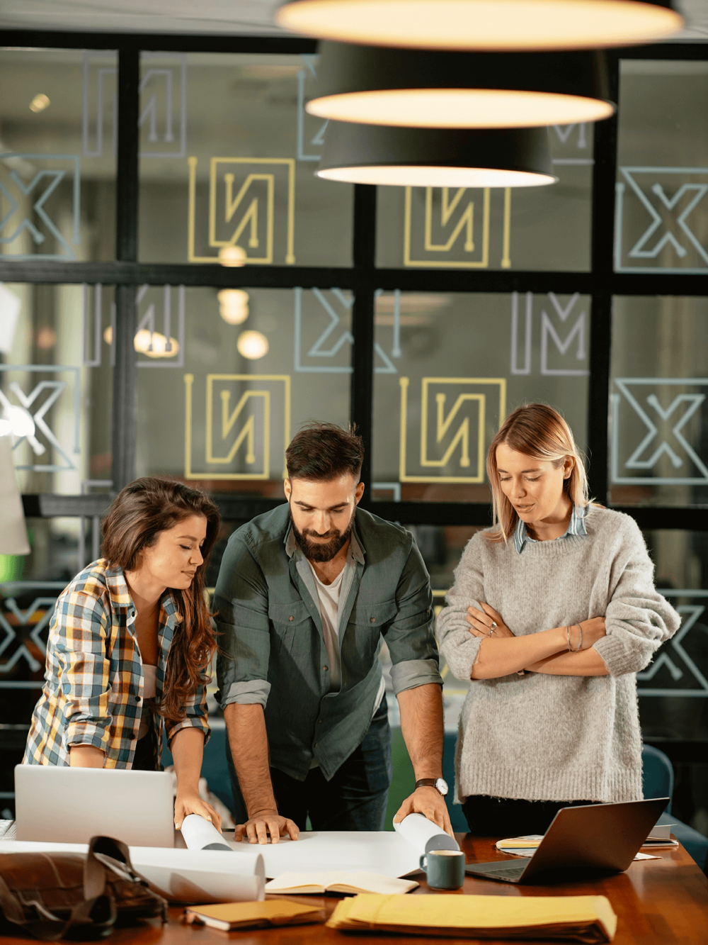 Three coworkers examining blueprints together at a table in a modern office setting.