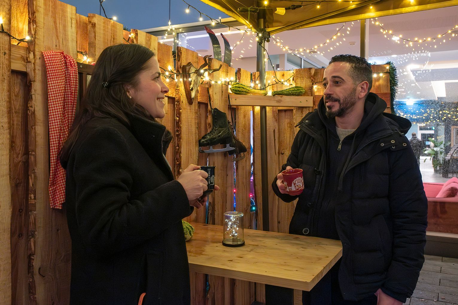 Zwei Personen unterhalten sich an der geschmückten Almhütte im Freien mit Lichterketten und Holzwänden.