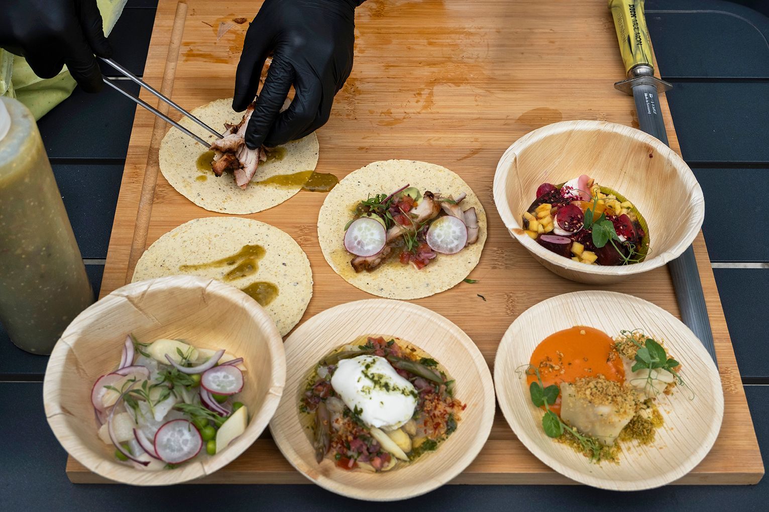 Hände richten bunte Tacos auf einem Holzbrett an, dazu sechs kleine Schälchen mit Toppings.