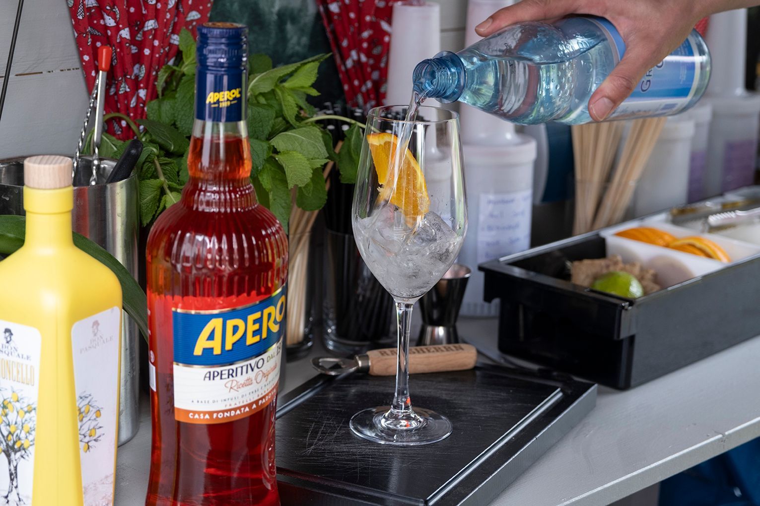 Ein Barkeeper gießt Wasser in ein Cocktailglas neben Aperol und Mixgetränken an einer Bar.