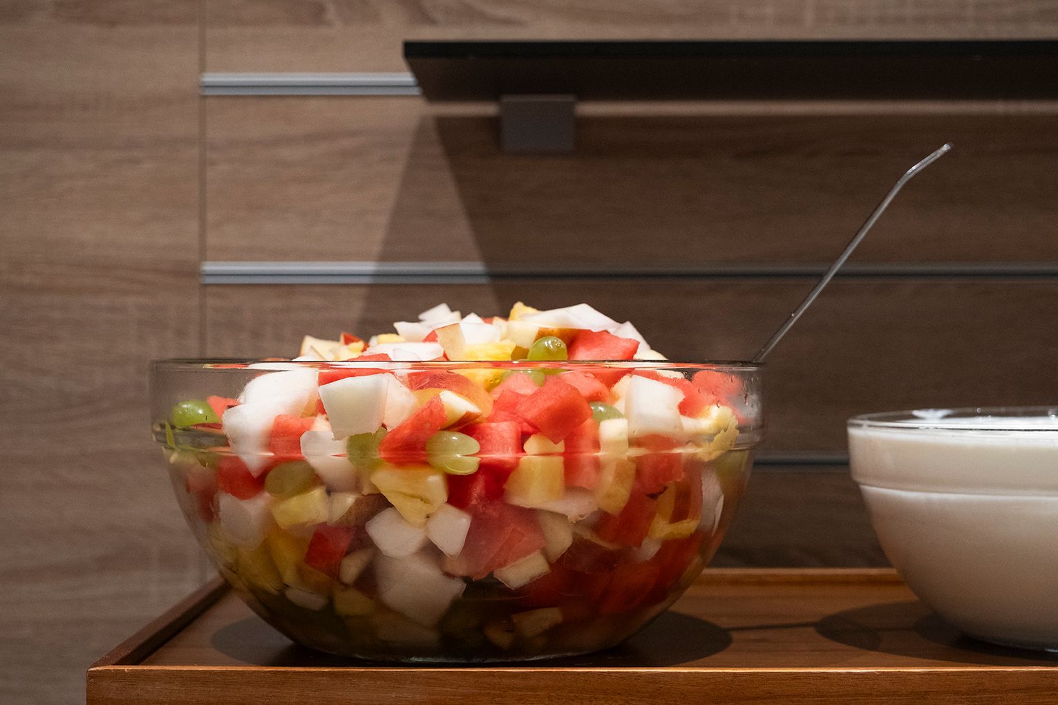 Eine Schüssel mit gehacktem Obstsalat und ein Löffel daneben, auf einer Holztheke.