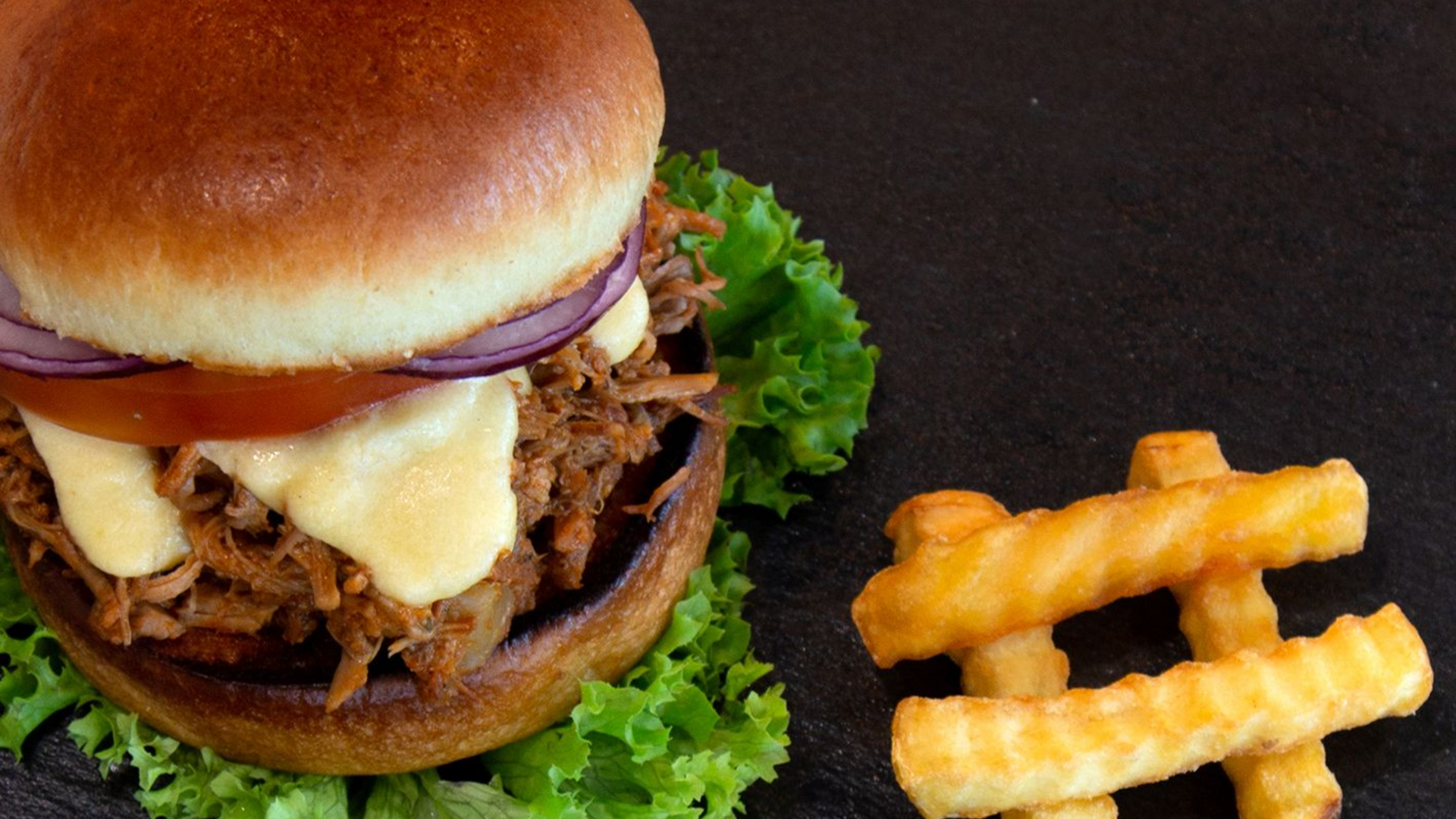 Burger mit geschmolzenem Käse und Salat auf einem dunklen Teller, dazu Wellenschnittpommes.