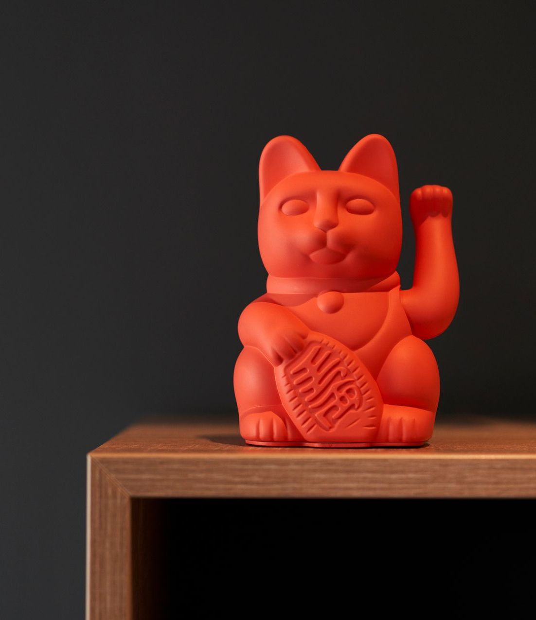 Red Maneki Neko cat figurine raising its paw, sitting on a wooden shelf against a dark gray background.