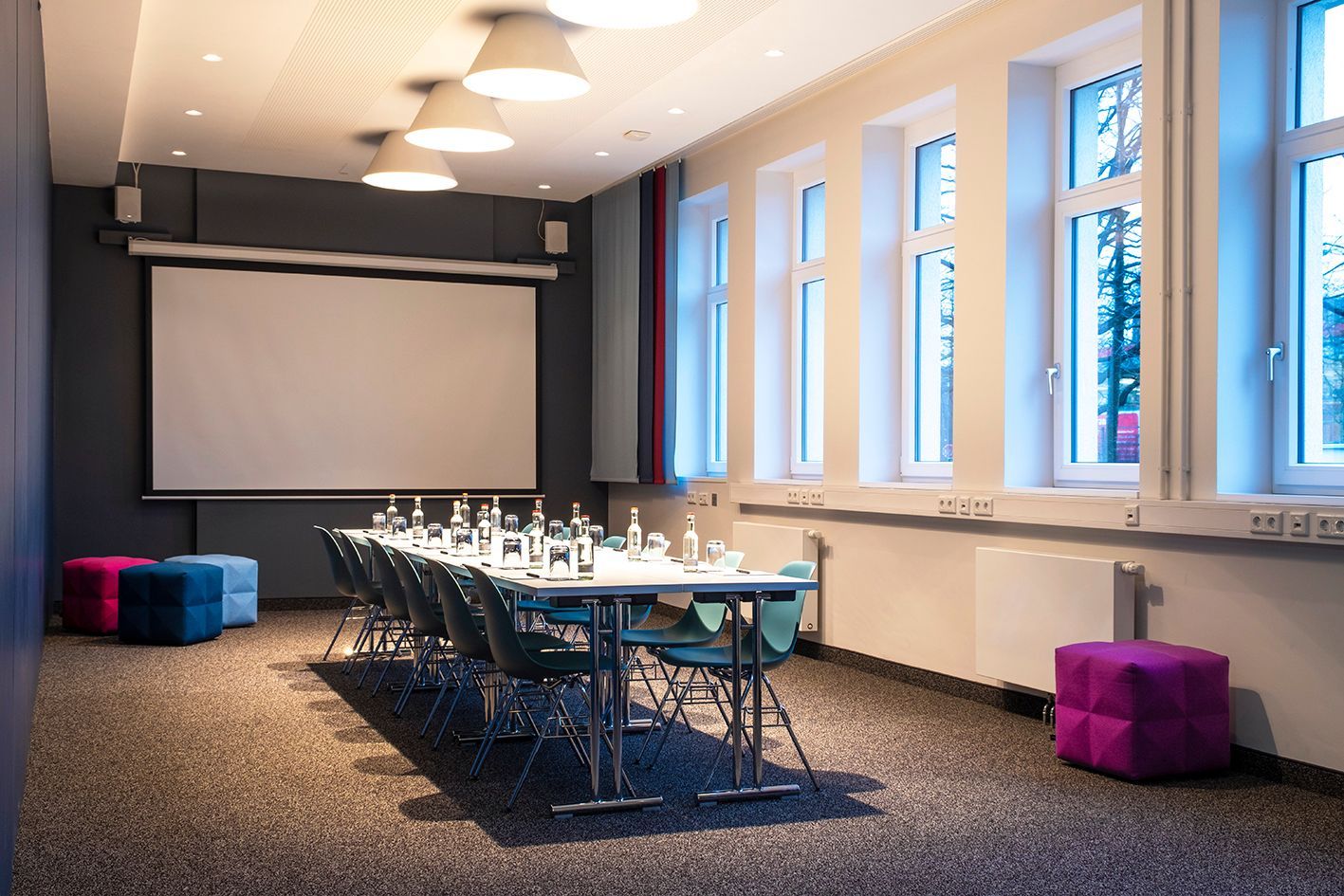 Study III Meetingraum mit langem Tisch, Stühlen, Projektionsleinwand und hellen Fenstern neben violetten Hockern.