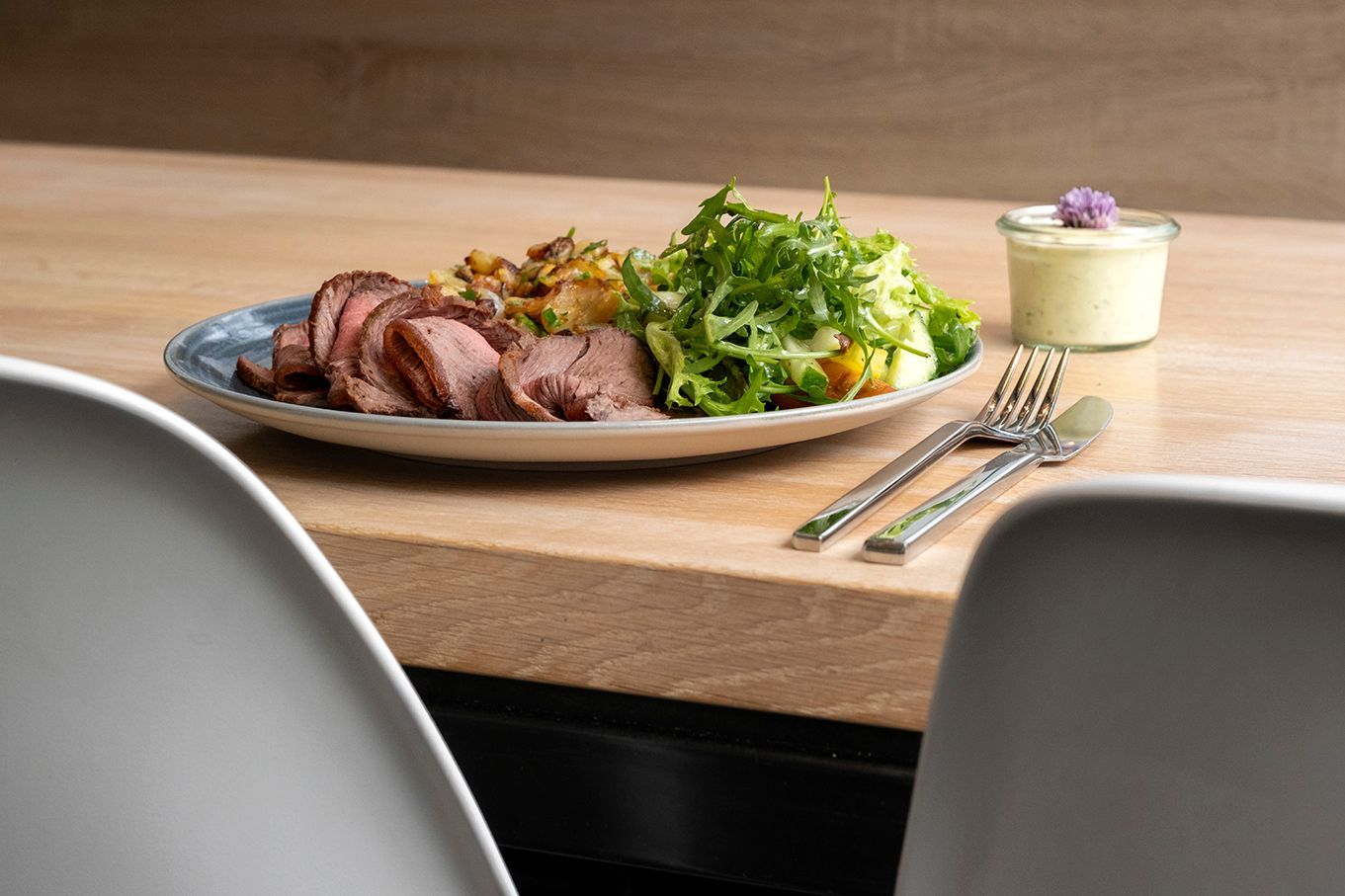 Ein auf einem Holztisch angerichteter Braten mit Gemüse, flankiert von zwei weißen Stühlen und Besteck.