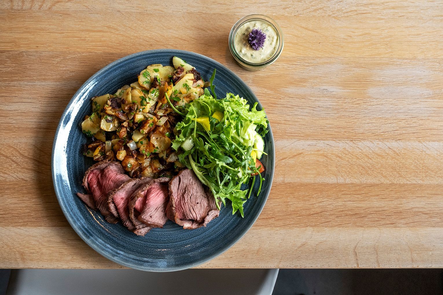 Ein Roastbeef mit Bratkartoffeln und Salat auf einem blauen Teller neben einem kleinen Saucenschälchen auf einem Holztisch.