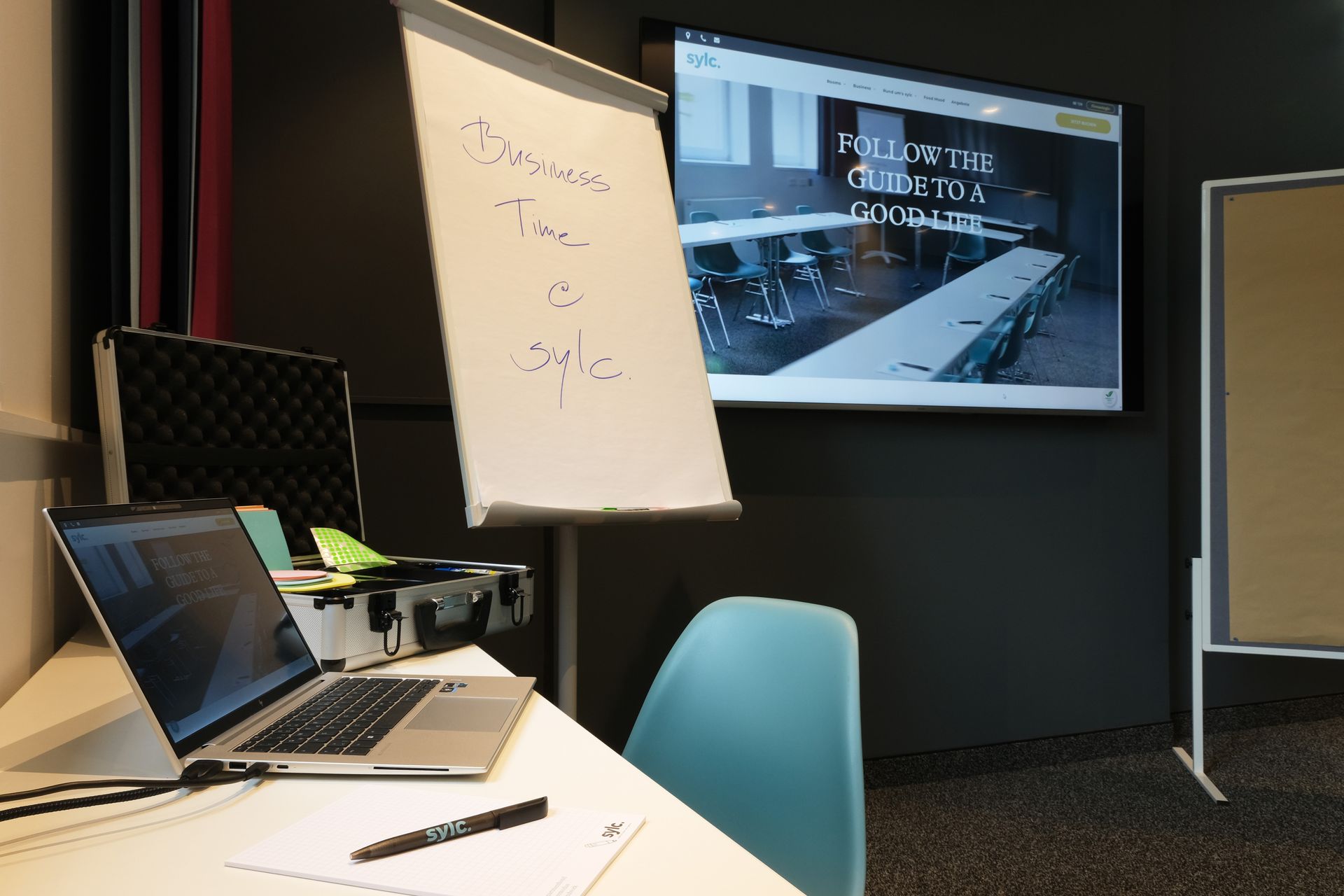 Ein Laptop und ein Whiteboard im Konferenzraum mit einem Bildschirm. Vor dem Tisch steht ein Stuhl.