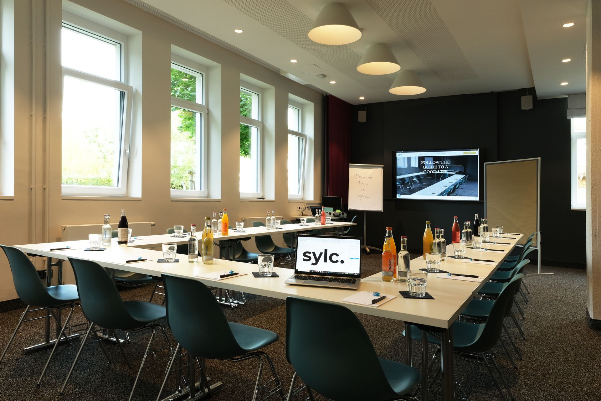 Konferenzraum mit U-förmigem Tisch, Stühlen, Getränken und einer Leinwand. Fenster und neutrale Wände.