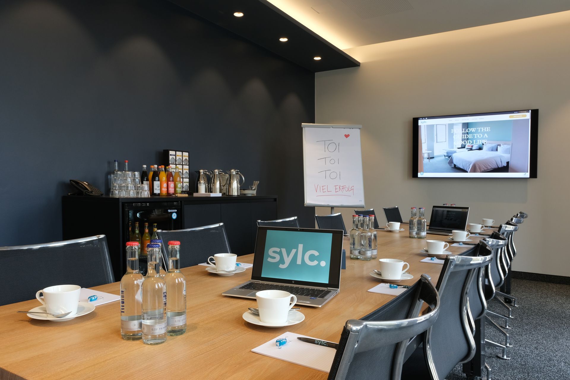 Ein Konferenzraum mit einem für ein Meeting gedeckten Tisch mit Bildschirm, Erfrischungen, Laptops und Flipchart.