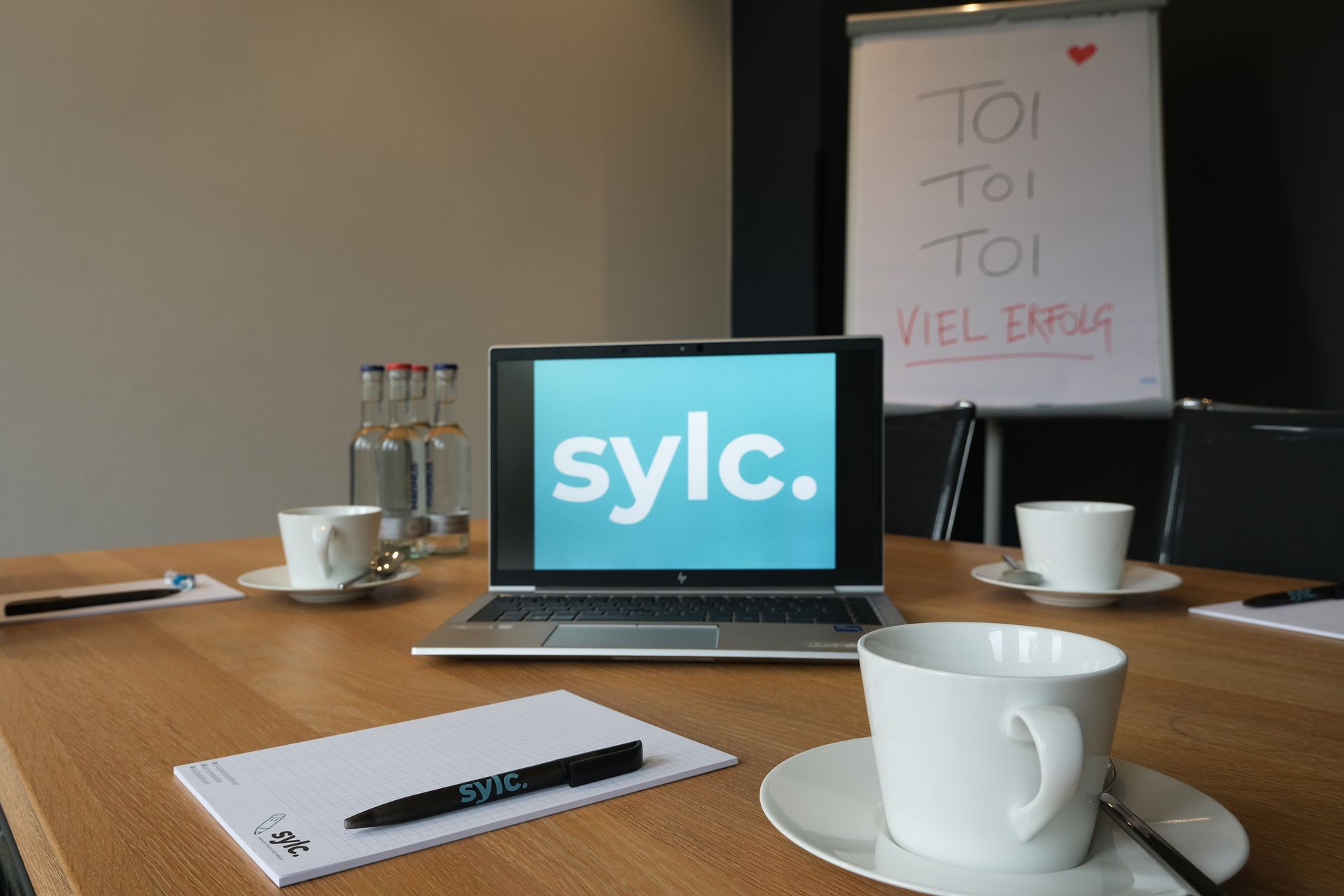 Laptop auf einem Konferenztisch mit Kaffeetassen, Wasserflaschen und Notizblock; im Hintergrund ein Whiteboard