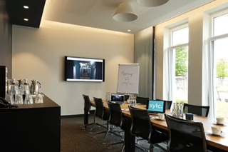 Ein Konferenzraum mit einem langen Holztisch, Stühlen und einem Whiteboard und Fernseher. Im Vordergrund steht eine Theke