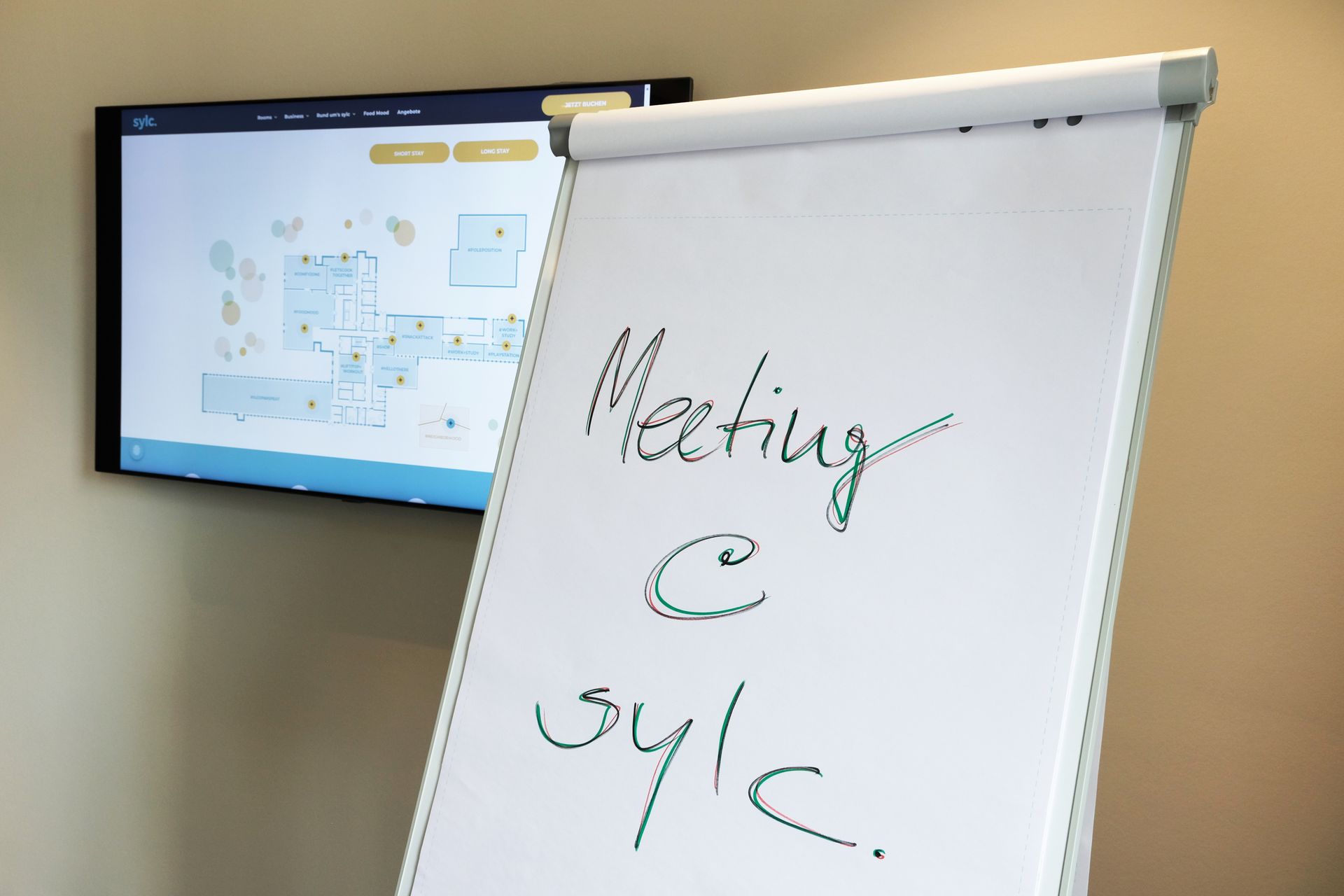 Flipchart mit der Aufschrift „Meeting @ sylc“ vor einem Bildschirm mit einem Gebäudeplan.