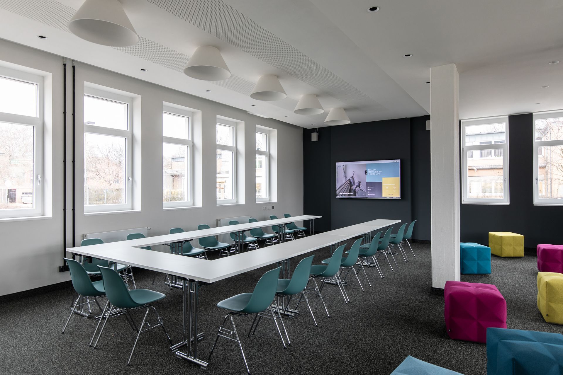 Ein moderner Konferenzraum mit einem U-förmigen Tisch, Stühlen und Ottomanen. An einer Wand ist ein Bildschirm angebracht.