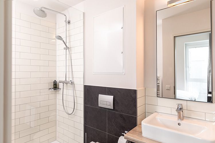 Badezimmer mit Fliesendusche und einem  Bereich mit Toilettenspülung. Daneben befinden sich ein Spiegel und ein Waschbecken.