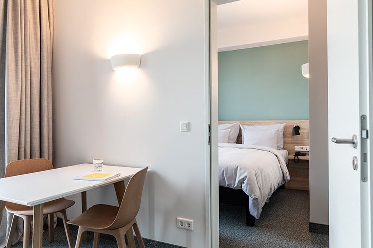 A modern hotel room with a large bed, a red ottoman, and a small side table. The wall behind the bed is mint green.