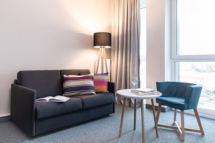 Apartment mit grauem Sofa, Lampe, kleinem Tisch und einem blauen Stuhl.