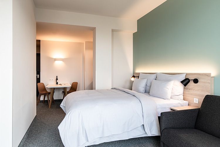 Modernes Hotelzimmer mit Bett, Sofa und Essbereich. Die Wände sind in hellen Farben mit einem blaugrünen Akzent gehalten.
