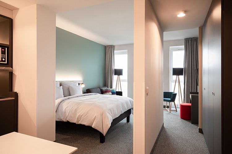 A modern hotel room with a large bed, a red ottoman, and a small side table. The wall behind the bed is mint green.