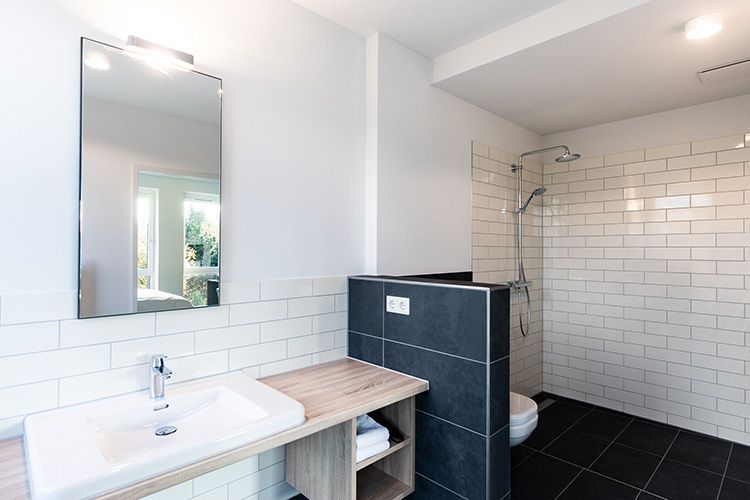 Modernes Badezimmer mit weißen Fliesen, schwarzem Boden und Glasdusche. Ein Waschtisch mit großem Spiegel ist sichtbar.