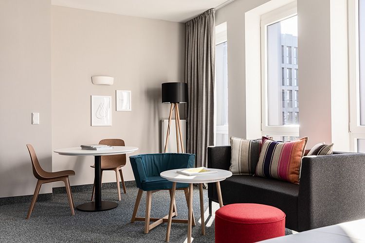 Apartment mit grauem Sofa, türkisfarbenem Sessel, rundem Tisch, Holzboden, Kunstwerken und Fenster.