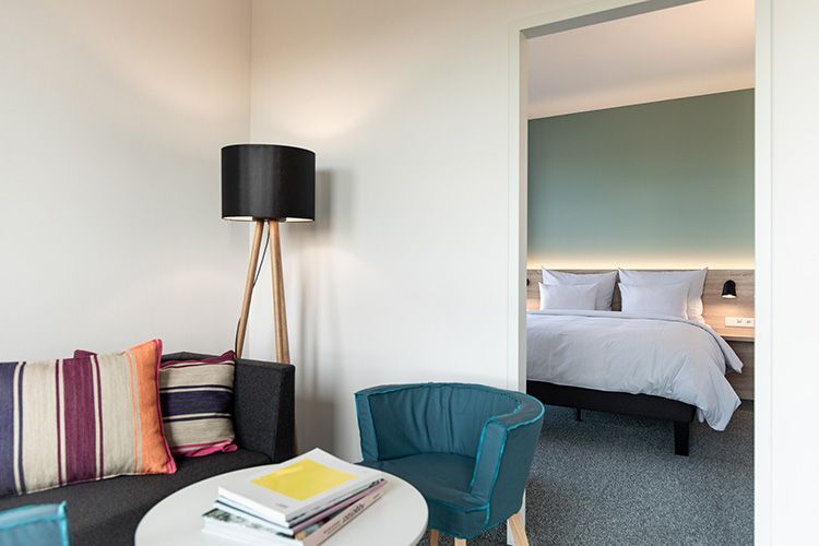 A modern hotel room with a large bed, a red ottoman, and a small side table. The wall behind the bed is mint green.