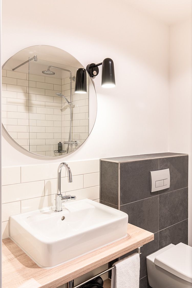 Ein Badezimmer mit einem Spiegel über einem Waschbecken und einer Leuchte. Hinter der Toilette ist eine graue Wand sichtbar.