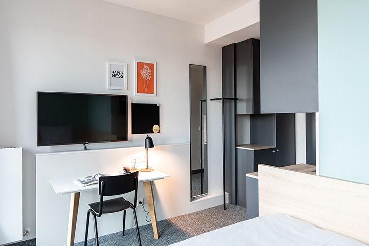 Modern hotel room with desk, chair, TV, and wall-mounted storage. Gray walls, light wood, and a minimalist design.
