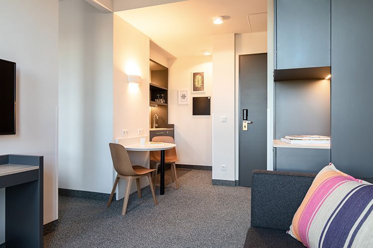 Ein kleines modernes Hotelzimmer mit einem Tisch, Stühlen und einer kleinen Küchenzeile im Hintergrund.