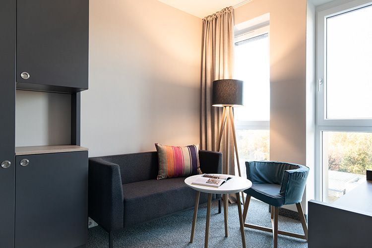 A modern hotel room with a large bed, a red ottoman, and a small side table. The wall behind the bed is mint green.