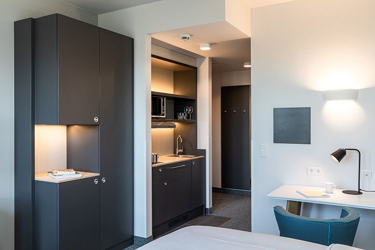 Apartment mit Kochnische und Schreibtisch, ausgestattet mit grauen Schränken, weißen Wänden und einem blauen Stuhl.