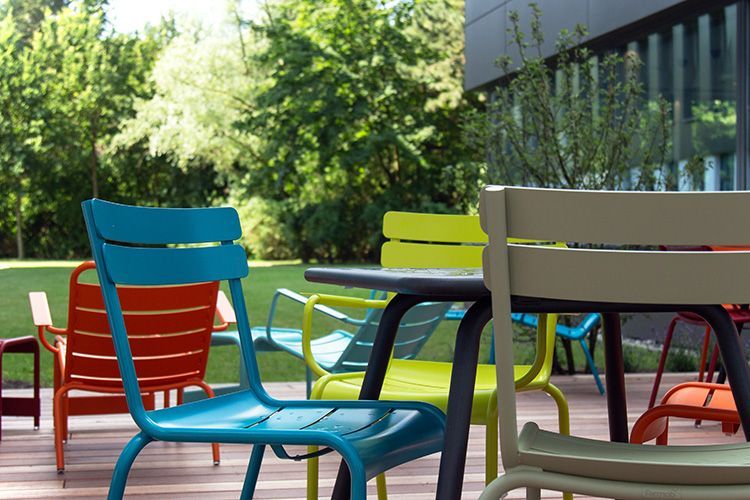 Bunte Metallstühle auf einer Terrasse im Freien, im Hintergrund Bäume und ein Gebäude.