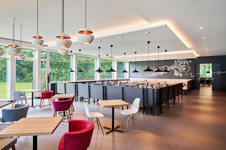 Helles Restaurant-Interieur mit Tischen, Stühlen und Hängelampen. Große Fenster blicken auf einen grünen Rasen.