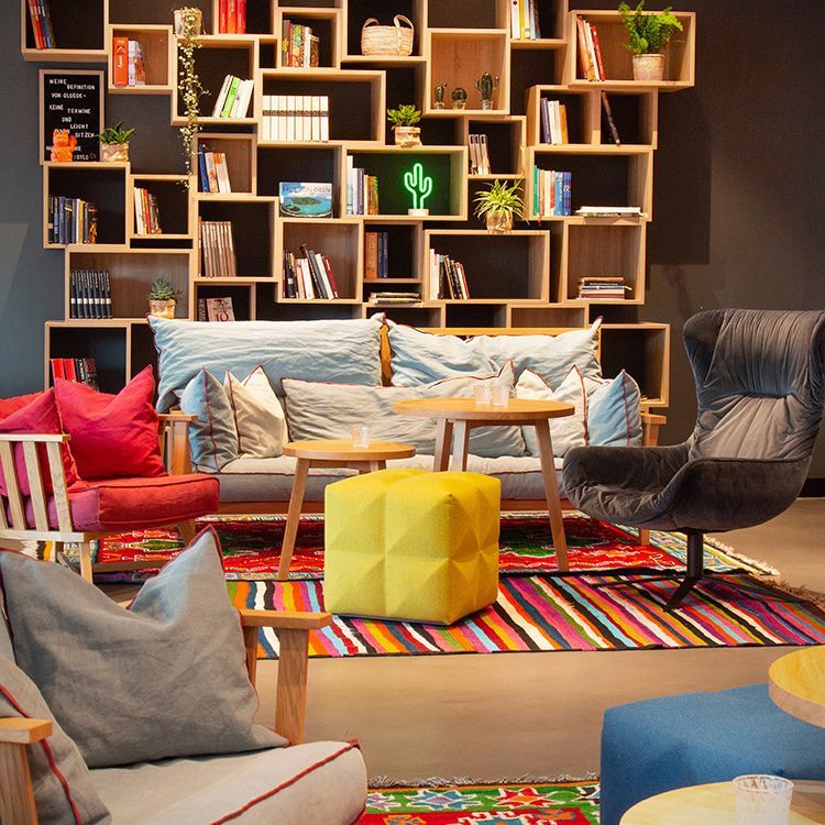 LIBRARY BAR mit Bücherregalen, Sofas, Sesseln und einem leuchtenden, geometrischen Hocker auf einem gestreiften Teppich.