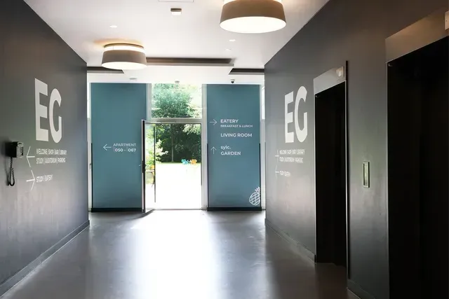 SICON Hospitality | Head of Sales | Nina Langen Flur mit dunklen Wänden, weißem „EG“-Logo, Aufzügen und einer Tür mit hellem Blick nach draußen.