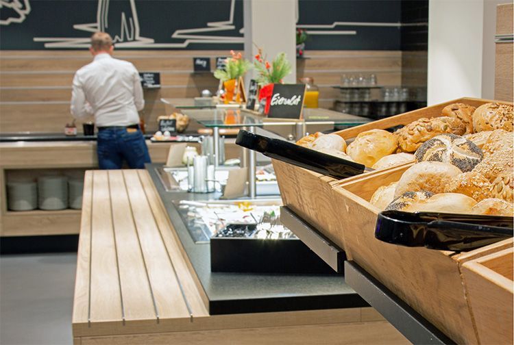 Ein Frühstücksbuffet mit verschiedenen Brotsorten. An einer Essensstation ist eine Person in einem weißen Hemd zu sehen.