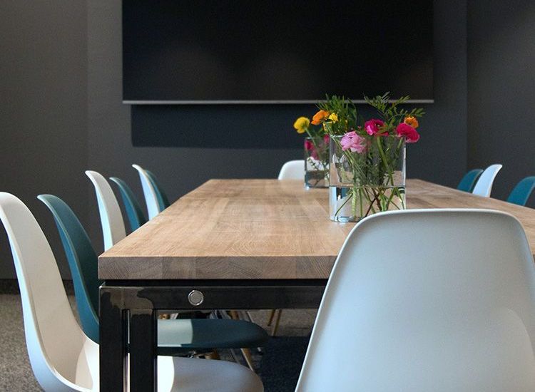 Meeting room with long wooden table, white and teal chairs, flowers in vases, and a dark wall.