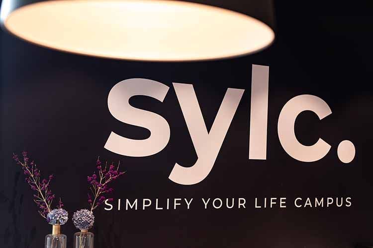 sylc. Apartmenthotel | Rezeption Zwei Frauen sitzen an einem an einer Rezeption vor einem Schild mit der Aufschrift „SYLC“.
