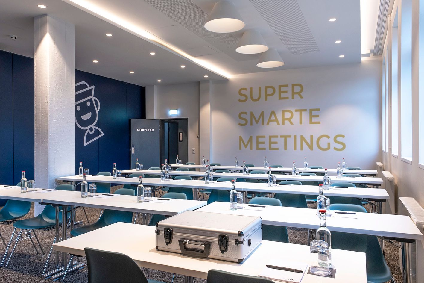 Study Lab Meetingraum mit Reihen weißer Tische, blauen Stühlen und dem Schriftzug „SUPER SMARTE MEETINGS“ an der Wand.
