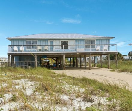 Elevated beach house on stilts with pale blue siding, sandy foreground, and clear blue sky