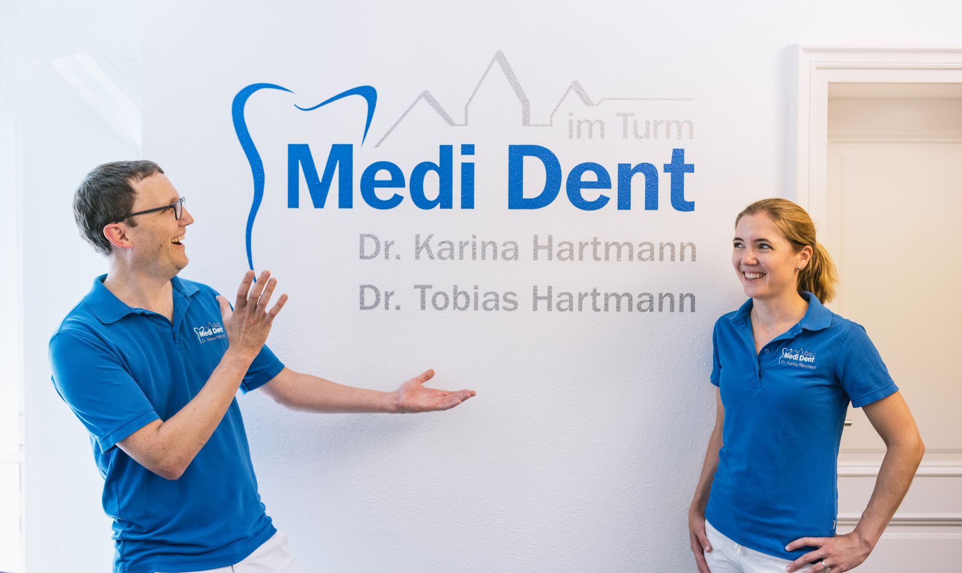 Dr. Tobias Hartmann und Dr. Karina Hartmann präsentieren das MediDent-im-Turm-Logo an der Wand ihrer Praxis
