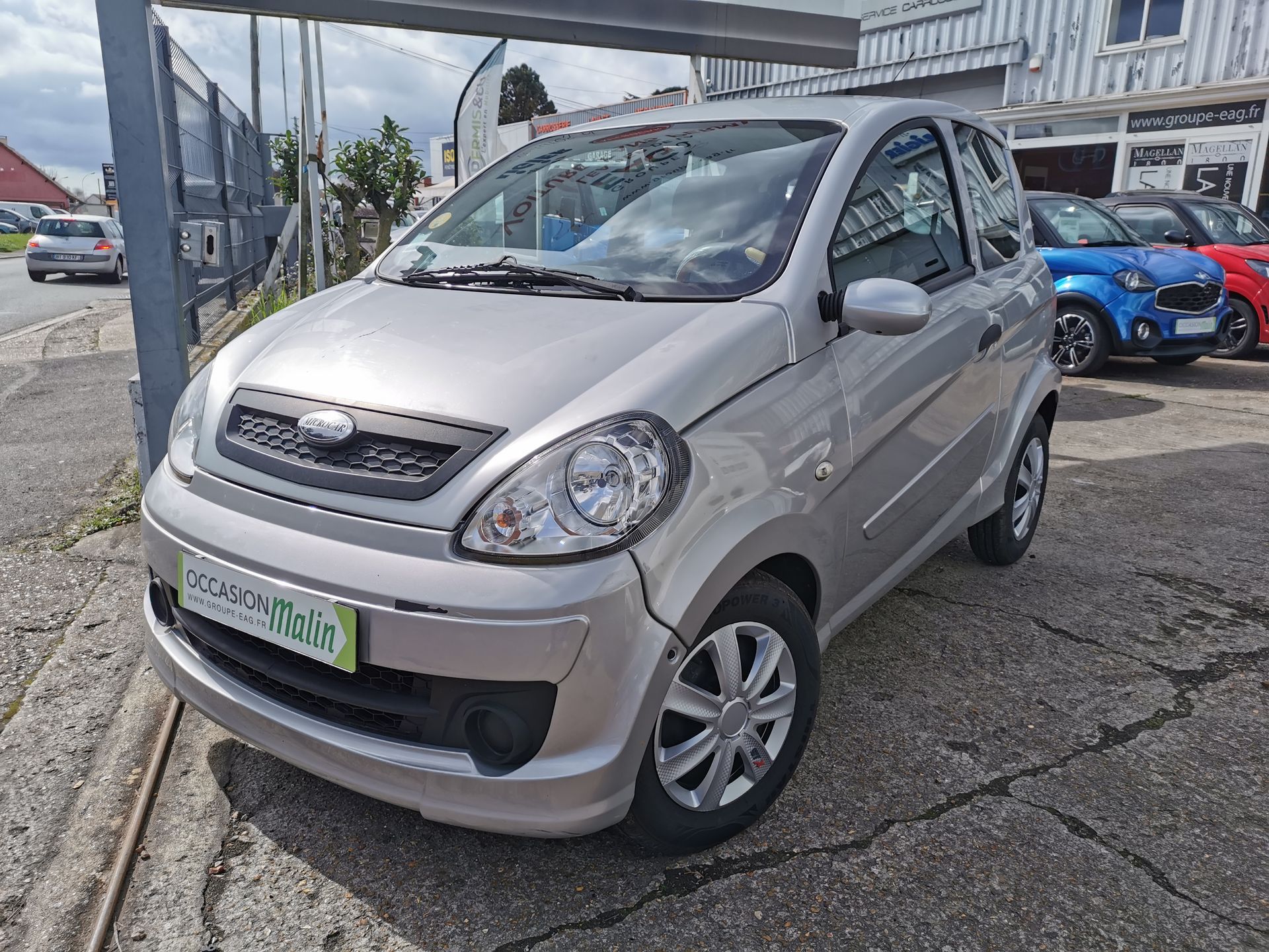 Voiture de ville Silver Aixam garée sur un parking goudronné, avec d'autres voitures en arrière-plan.