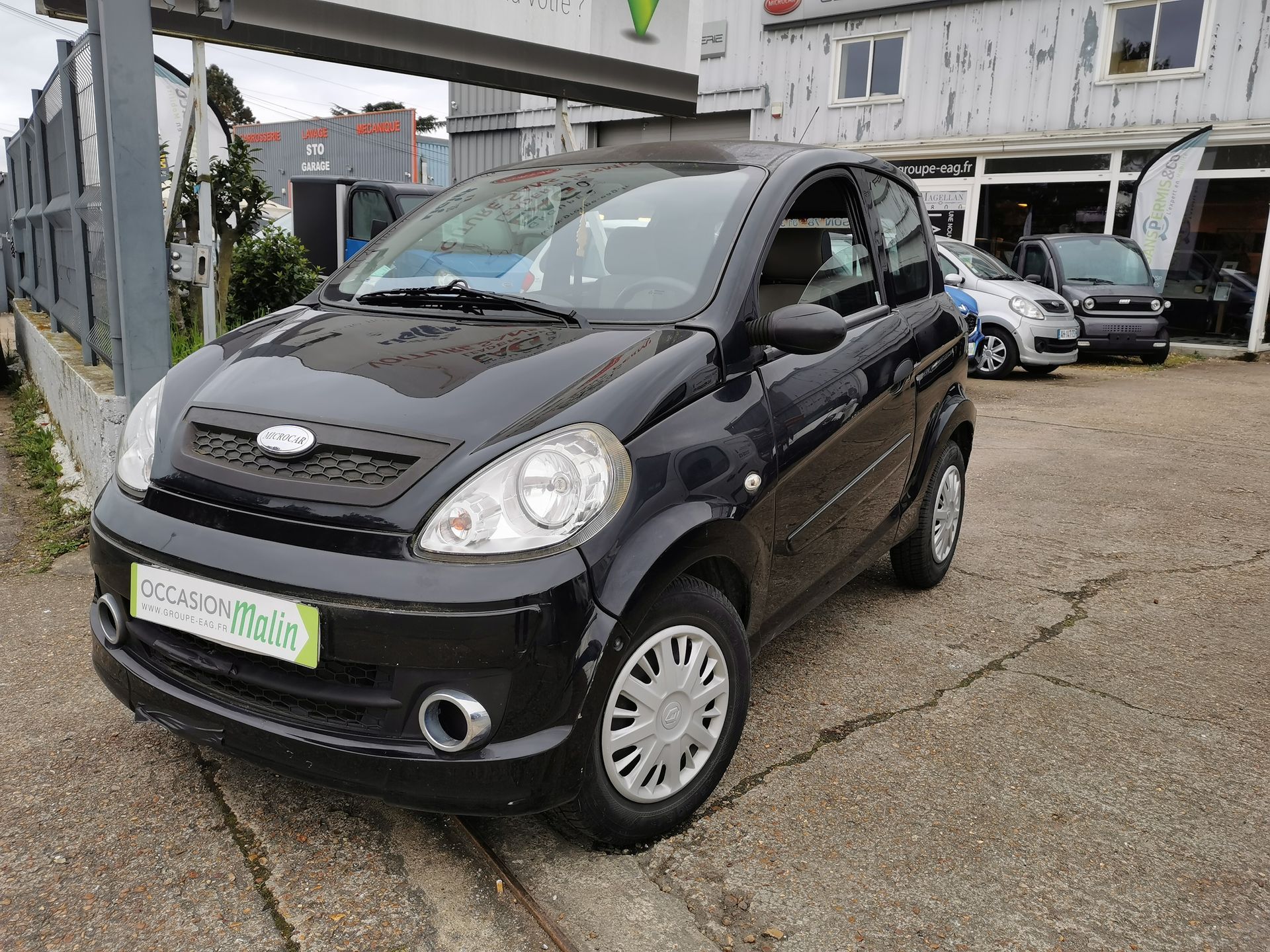 Voiture citadine noire Aixam garée sur du béton, devant une concession automobile.