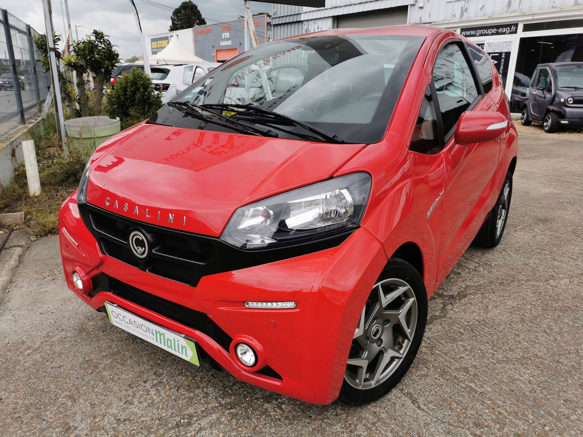 Voiture Aixam rouge garée sur un parking goudronné, avec une concession automobile en arrière-plan.