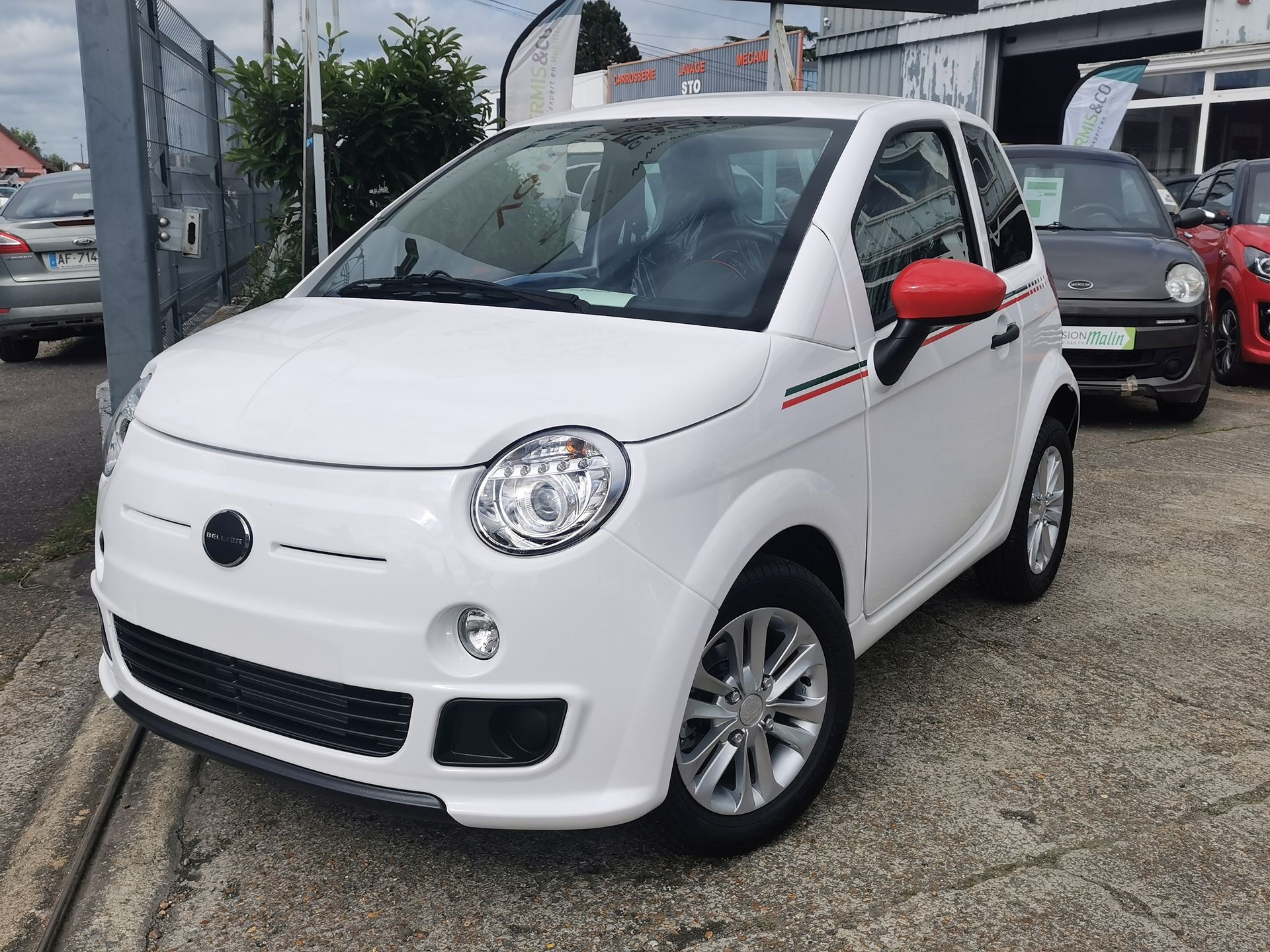 Voiture blanche deux portes avec rétroviseurs rouges et détails noirs, garée sur un parking.