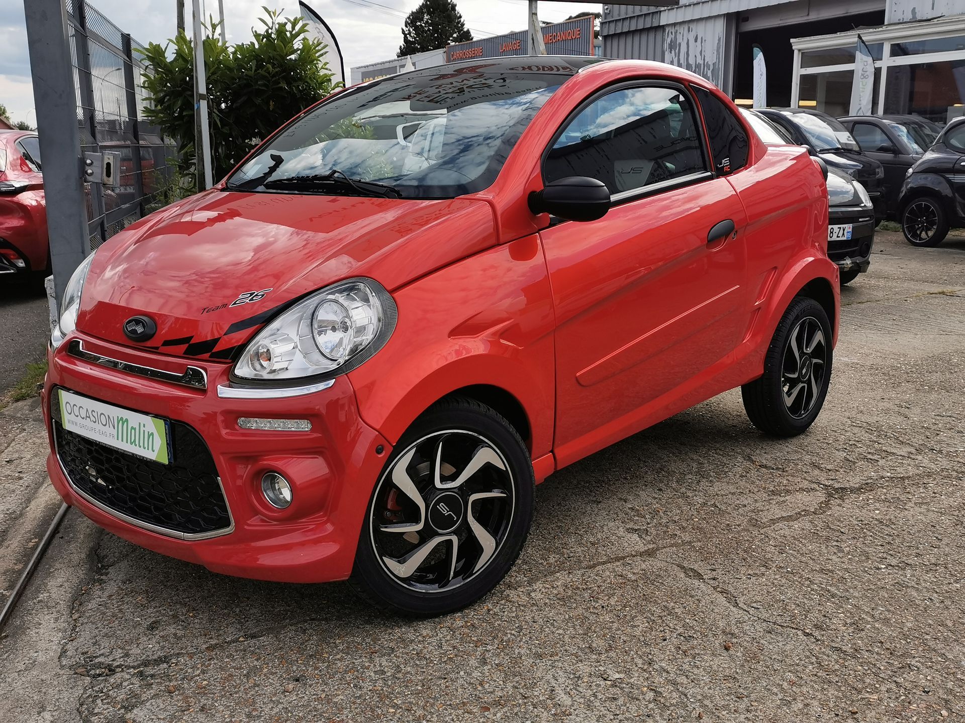 Une microvoiture rouge deux portes est garée dehors, avec des accents noirs, un toit noir et des roues noires et argentées, sur du gravier.