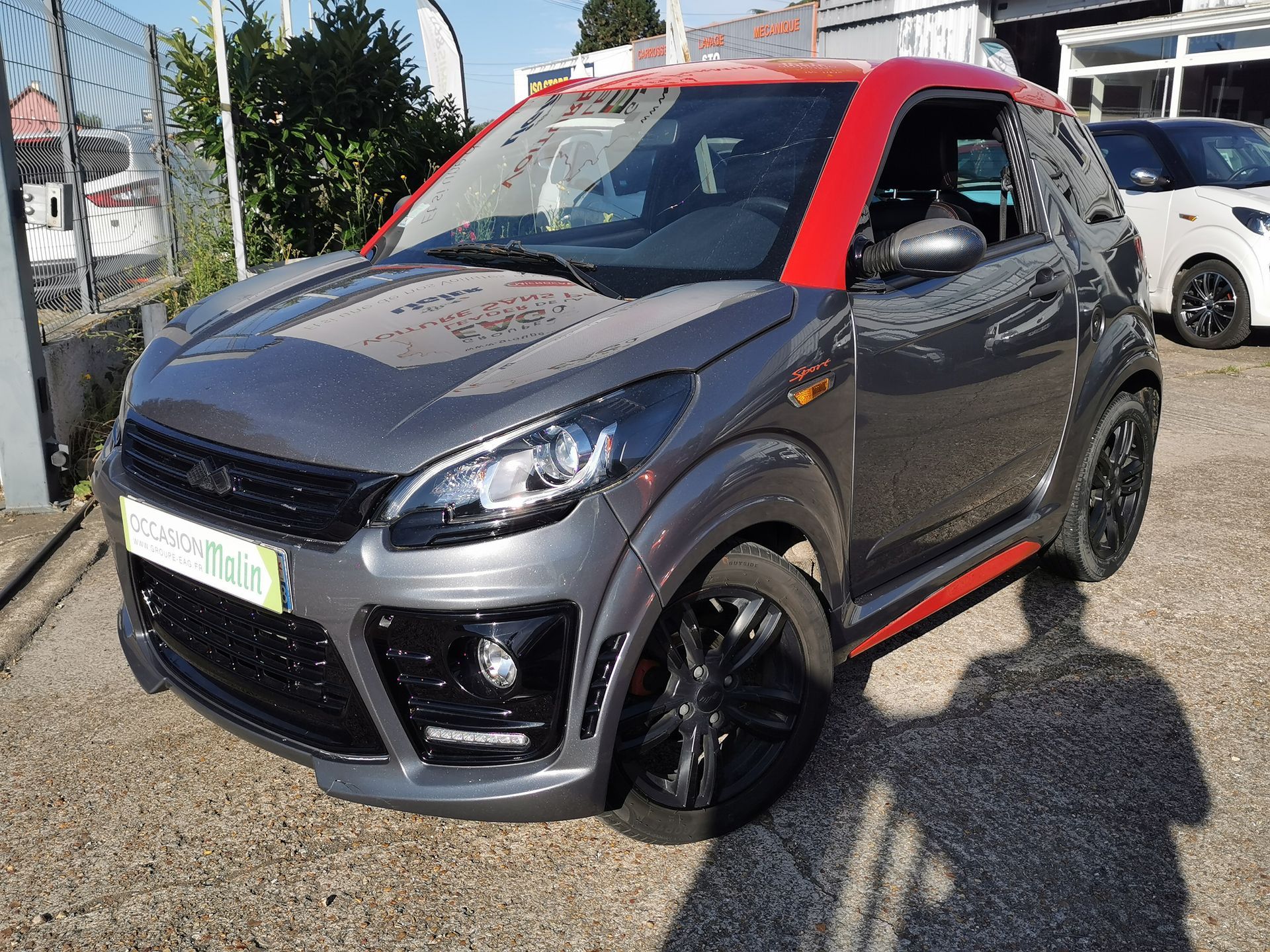 Voiture Aixam grise et rouge garée dehors par une journée ensoleillée.