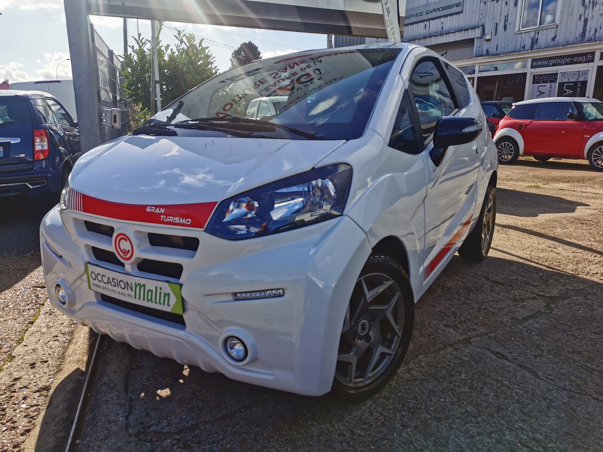 Une microvoiture blanche aux accents rouges est garée à l'extérieur, sous un ciel partiellement ensoleillé.