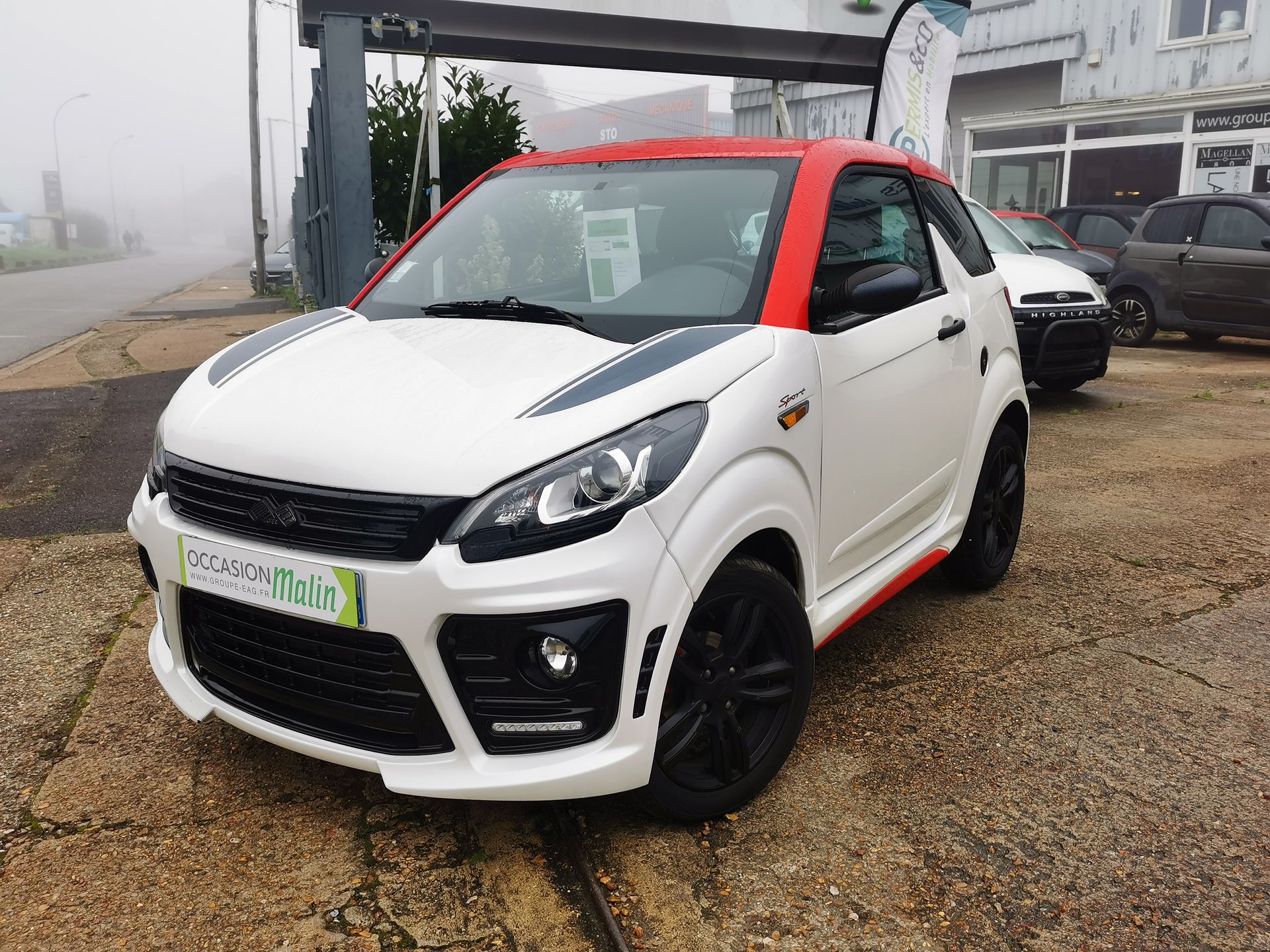 Voiture compacte blanche et rouge avec des touches de noir, garée devant un bâtiment par temps de brouillard.