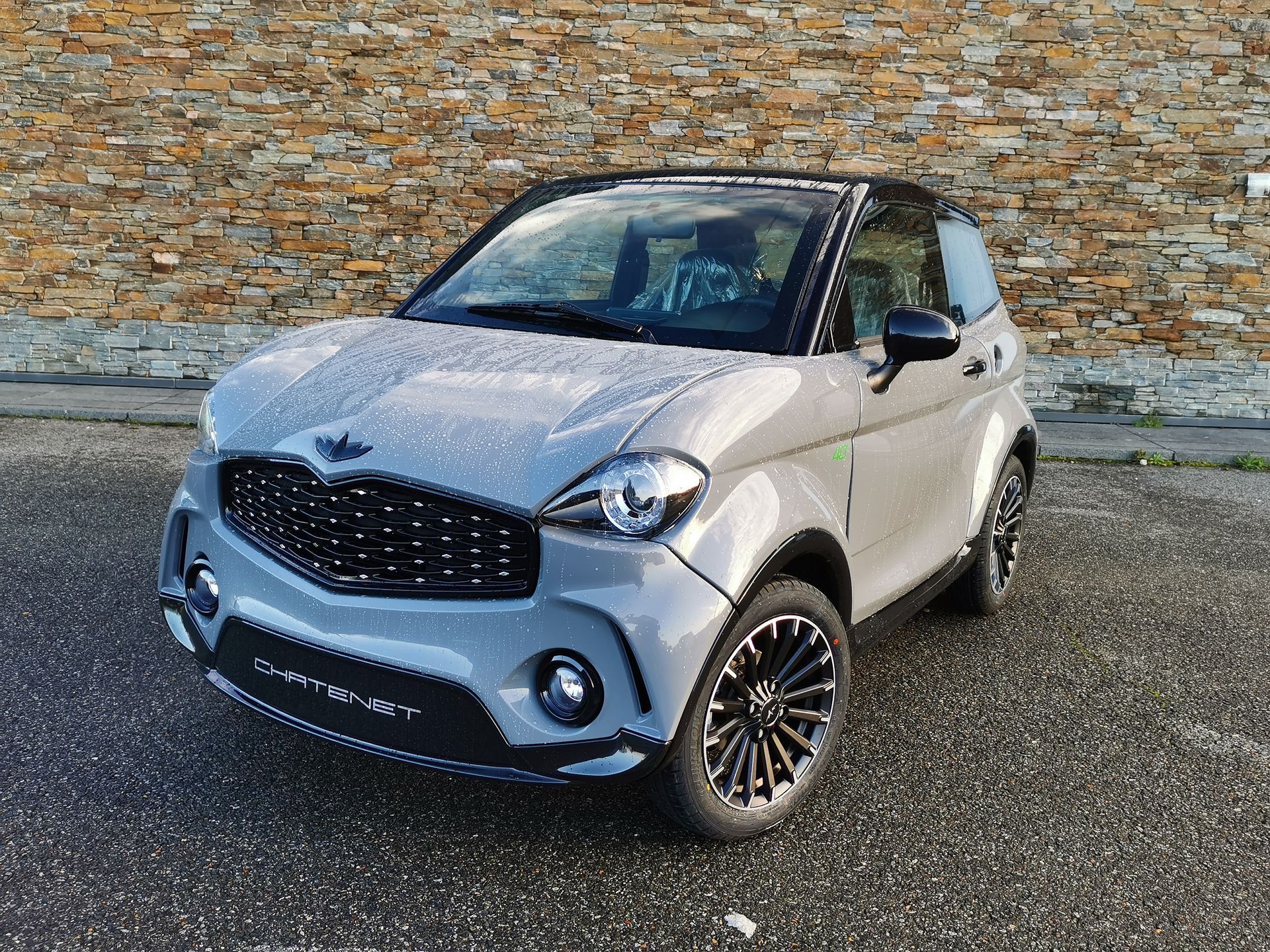 Voiture compacte deux portes grise et noire à jantes noires, garée devant un mur en pierre.