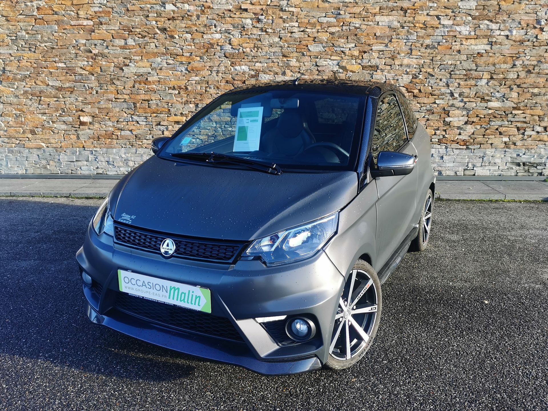 Coupé Aixam gris foncé garé devant un mur de pierre. Toit noir, jantes stylisées et enseigne verte du concessionnaire.